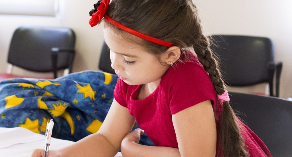 Une fillette au long cheveux et portant une boucle dans les cheveux est à une table et écrit dans un cahier.
