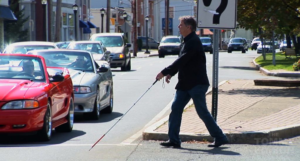 Un homme qui utilise une canne blanche traverse une rue. 