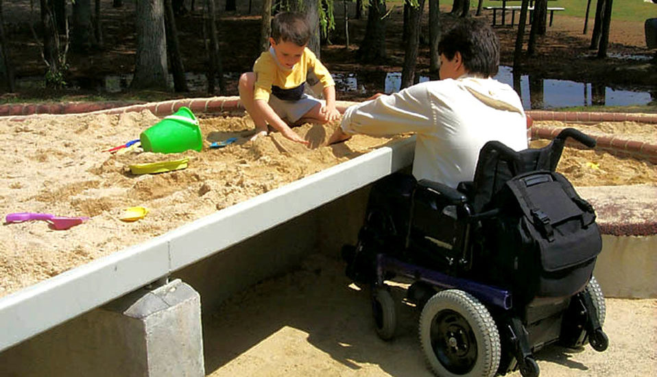 Un père en fauteuil roulant joue avec son petit garçon dans un bac à sable sur-élevé qui permet au fauteuil de se glisser dessous. 
