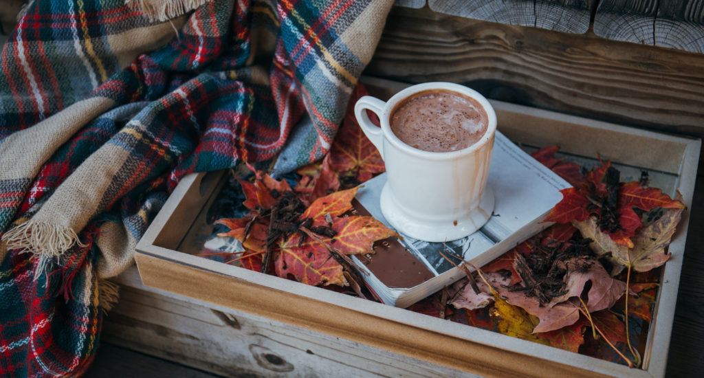 Une tasse de chocolat chaud déposée sur un livre, déposé sur des feuilles mortes, à côté d'un châle en laine à carreaux. 