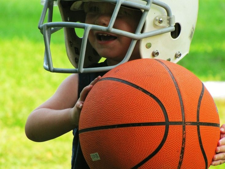 Un petit garçon porte un fros casque de football et tient dans sa main un ballon de basketball.