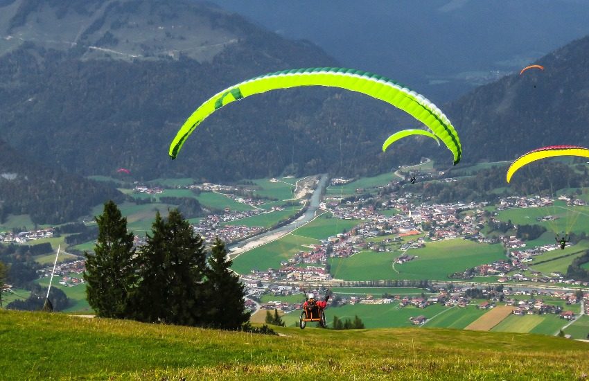 Une personne en fauteuil roulant fait du parapente, on voit un paysage de montagnes au loin. 