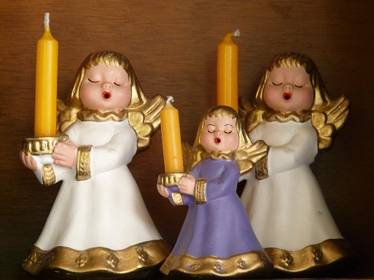 3  figurines d'anges, aux cheveux dorés et robes blanches, ont les yeux fermés  et la bouche ouverte comme s'ils chantaient. Ils tiennent de gros cierges dans leurs mains. 
