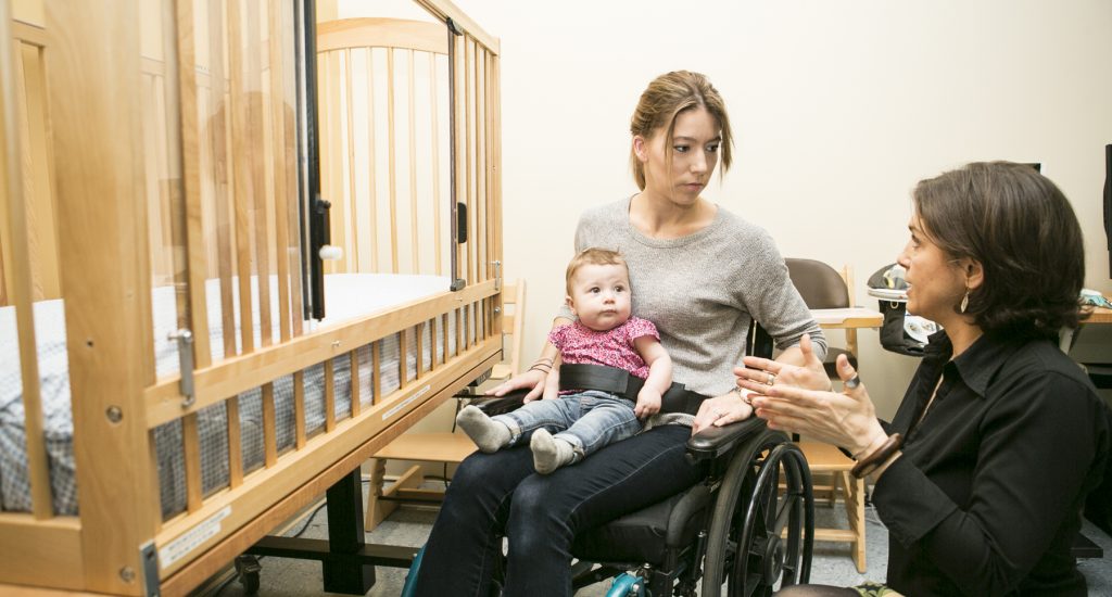 Une femme en fauteuil roulant a sur ses genoux une petit fille de moins d'un an, retenue par une ceinture. Agenouillée près d'elle, une femme lui explique comment utiliser un lit de bébé adapté . 
