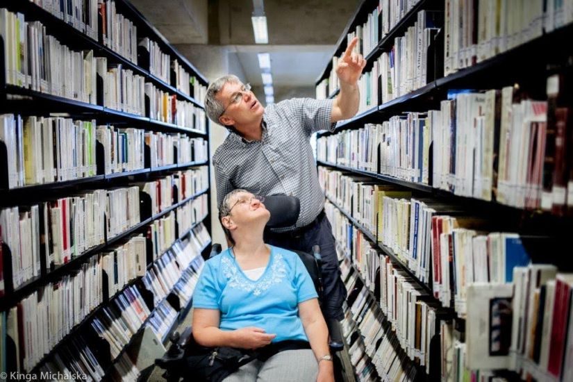 Un homme accompagne une femme en fauteuil roulant dans les rayons de livres de la Grande Bibliothèque. 