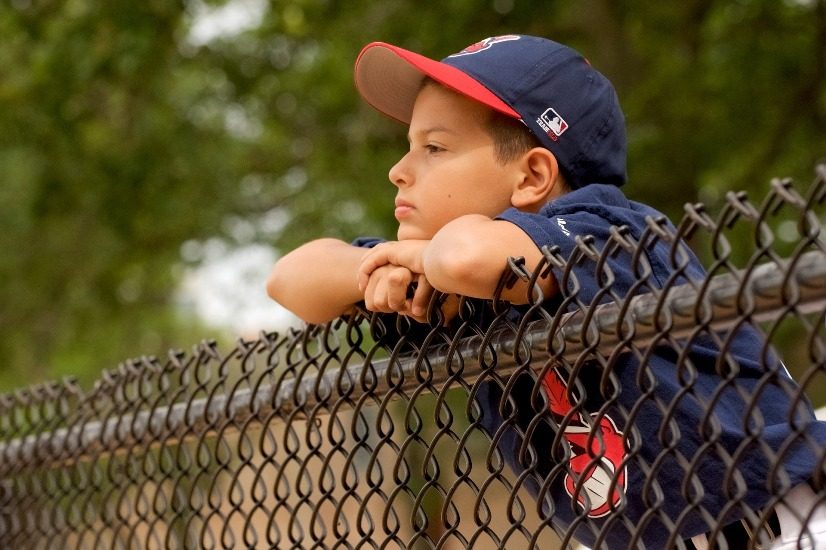 Un jeune garçon habillé pour jouer au baseball est appuyé sur une clôture métallique. Il regarde au loin et a un air sérieux et pensif. 
