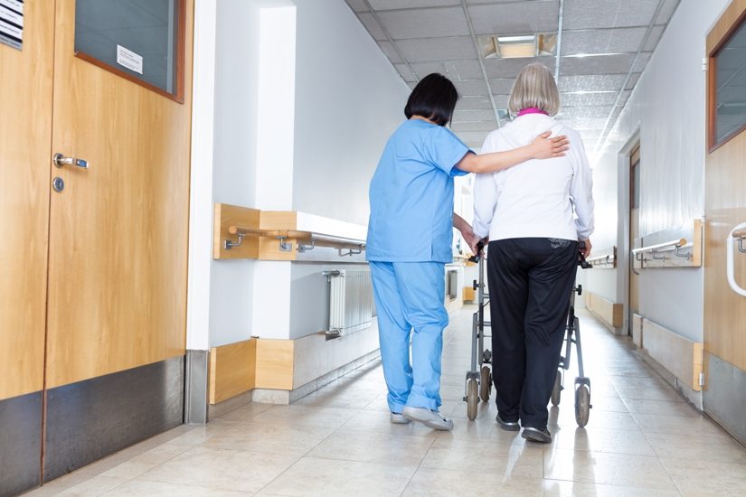 On voit de dos, dans un couloir, une préposée portant un uniforme bleu et une dame âgée qui utilise une marchette. 