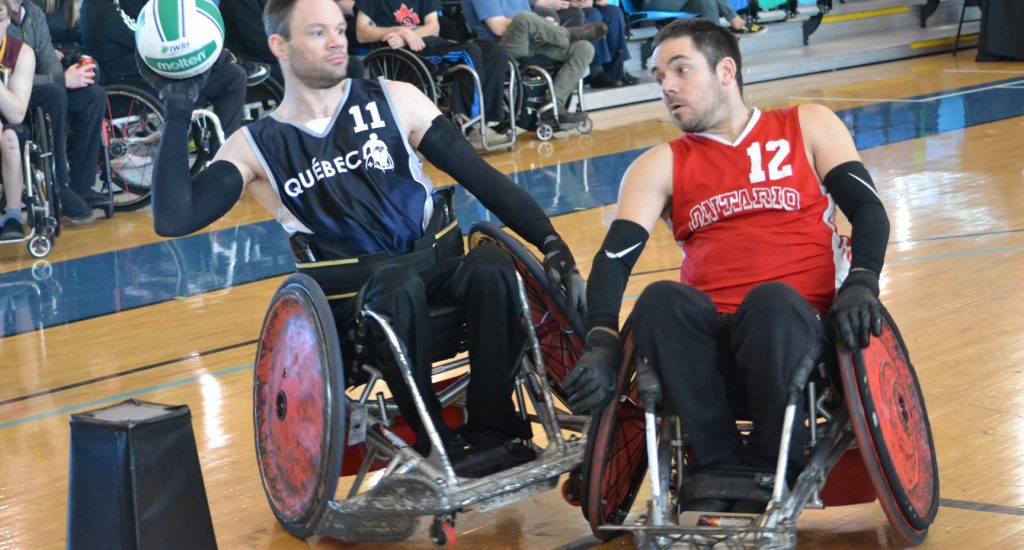 Fabien L.#11 Fabien Lavoie en plein action: en fauteuil roulant, il tient le ballon dans sa main tandis qu'un autre homme en fauteuil fonce dans sa direction. 