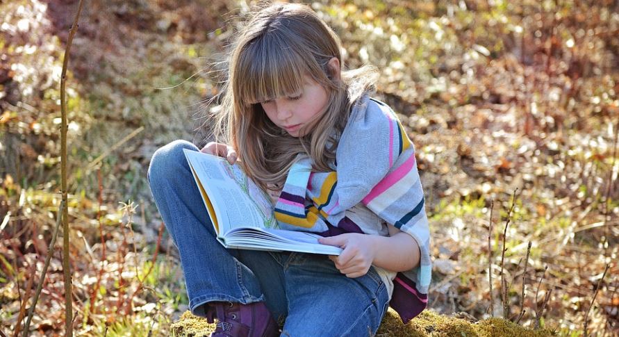 Une fillette blonde lit un livre, assise sur un sol jonché de feuilles mortes.