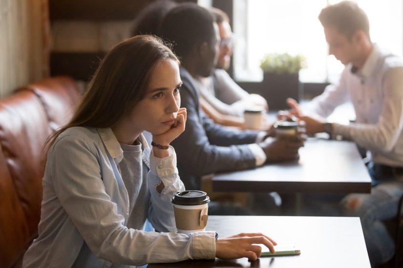 Une jeune femme aux cheveux bruns longs est seule à la table d'un café alors que plusieurs personnes discutent à la table voisine. 