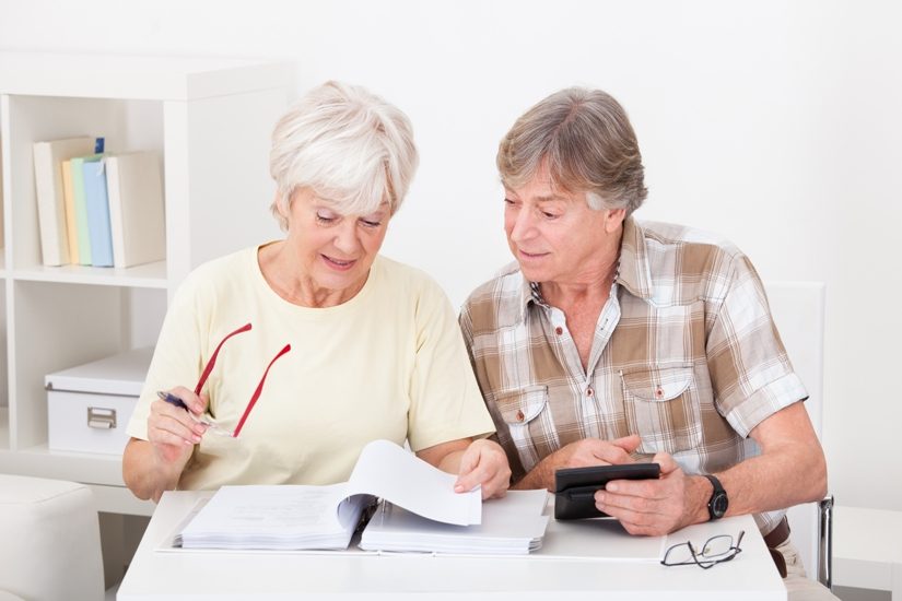 Un homme et une femmes à une table. Ils ont tous les deux les cheveux gris, la femme feuillette un document et l'homme a une calculatrice à la main. 