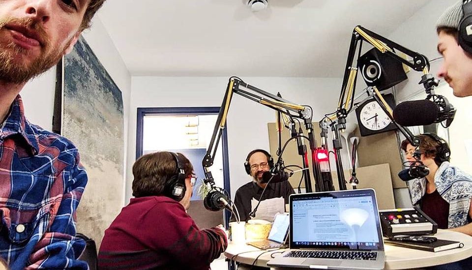 On voit l'équipe du balado en studio, avec micros, écouteurs et ordinateurs portables. Une lumière rouge indique que les micros sont allumés. On voit l'équipe du balado en studio, avec micros, écouteurs et ordinateurs portables. Une lumière rouge indique que les micros sont allumés.