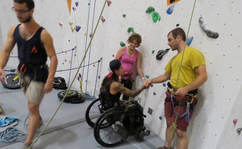 Devant un mur d'escalade, on voir deux personnes debout qui tendent la main à une femme en fauteuil roulant. Devant un mur d'escalade, on voir deux personnes debout qui tendent la main à une femme en fauteuil roulant.