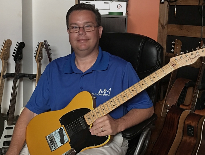 Tommy Théberge est assis sur une chose chaise de cuir noire et tient dans ses mains une guitare électrique. Il est photographié à l'école Musique O Max. 