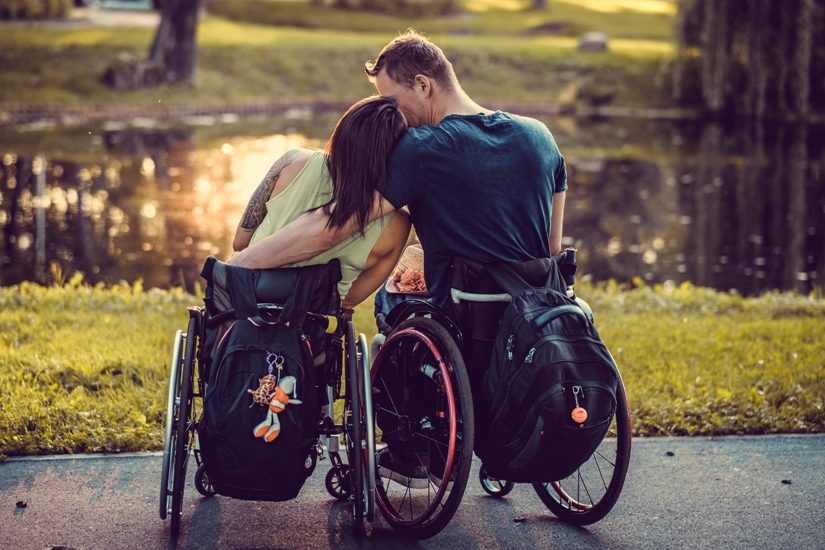 Sur la photo, un homme et une femme, tous deux en fauteuil roulant, devant un étang, dans un parc. La fille appuis ta tête sur l'épaule de l'homme. 
