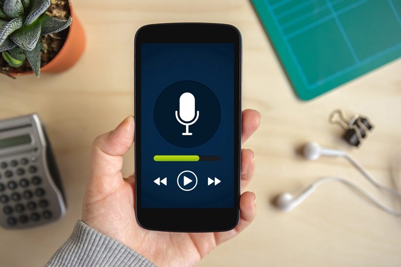 Au-dessus d’une table une main caucasienne tiens un téléphone intelligent. Tout prêt sur la table, on voit des écouteurs. Sur le téléphone, on voit un dessin de microphone, symbole usuel de la balladodiffusion. Au-dessus d’une table une main caucasienne tiens un téléphone intelligent. Tout prêt sur la table, on voit des écouteurs. Sur le téléphone, on voit un dessin de microphone, symbole usuel de la balladodiffusion.