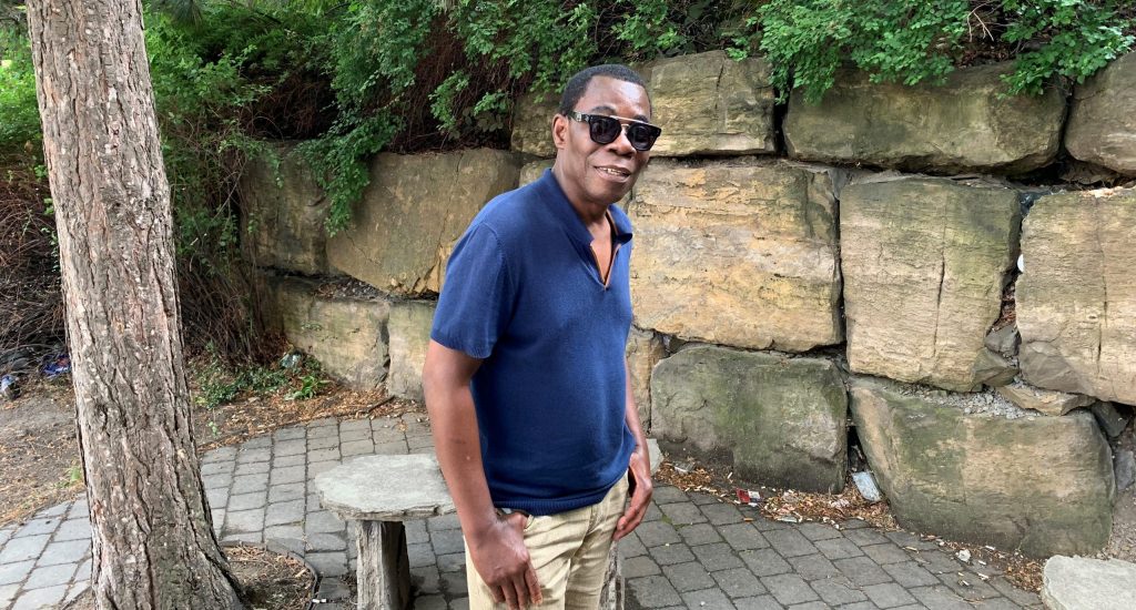 Fulgence Bla, devant un mur de pierre, avec un chandail bleu et des lunettes fumées Fulgence Bla, devant un mur de pierre, avec un chandail bleu et des lunettes fumées