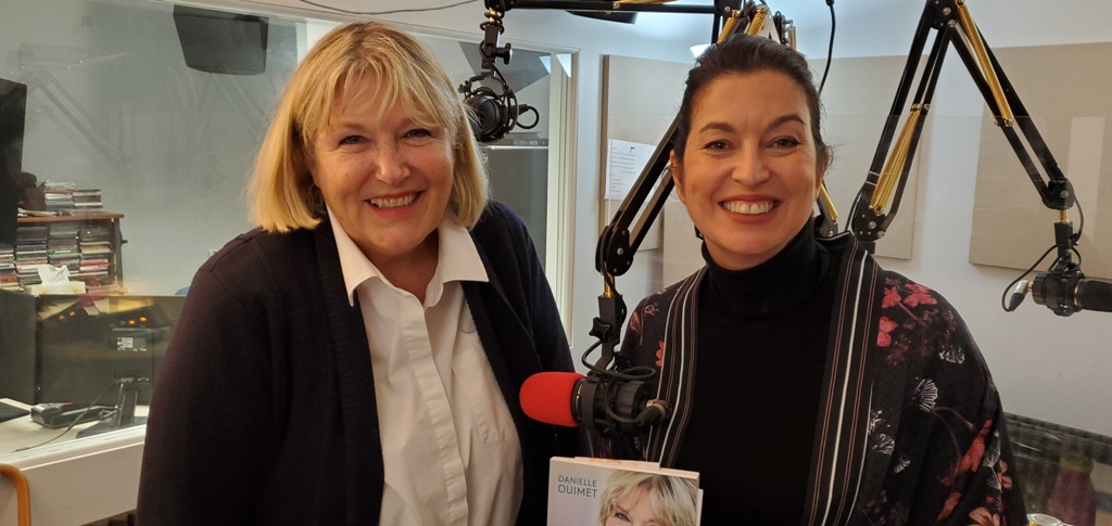 À gauche, Danielle Ouimet, une femme blonde. À droite, Chantal Dauray, une femme aux cheveux noirs. Elle sont en studio et Chantal tient dans ses mains la biographie de Mme Ouimet. À gauche, Danielle Ouimet, une femme blonde. À droite, Chantal Dauray, une femme aux cheveux noirs. Elle sont en studio et Chantal tient dans ses mains la biographie de Mme Ouimet.