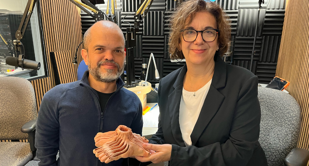 Hélène Denis pose en studio avec Matias Sierra, un homme de petite taille, tenant une sculpture en forme de pied et de volcan dans ses mains.