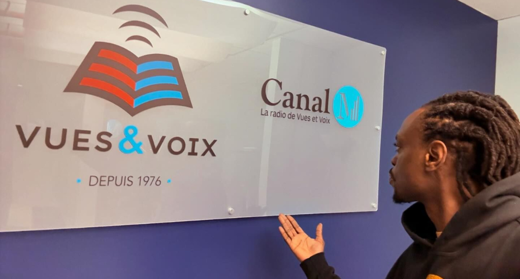 Ekinoxx pointe avec sa main l'enseigne «Vues et Voix / Canal M».