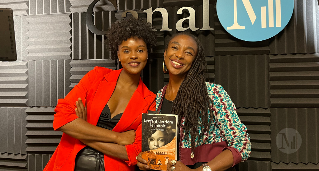 Keithy Antoine et Mariana Baldé posent debout en studio devant le logo de Canal M. Keithy tient le livre : «L'enfant derrière le miroir.»